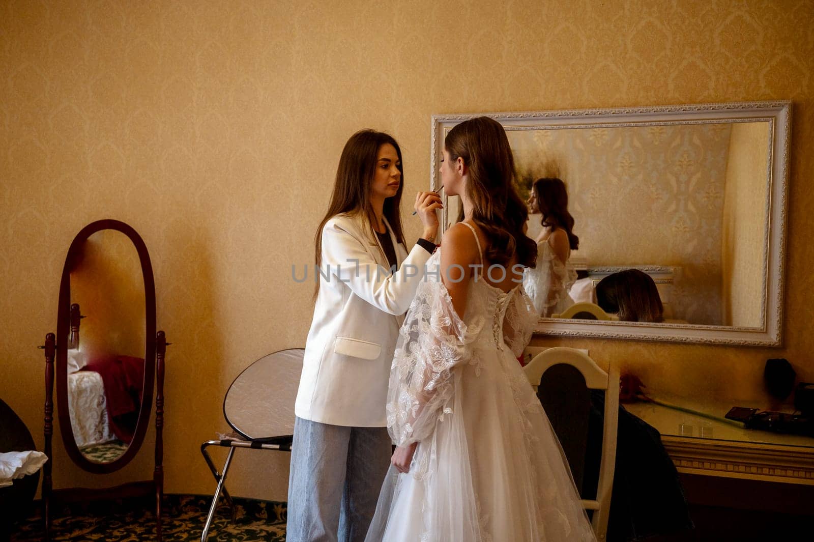 A stylist or makeup artist prepares a bride for a wedding indoors. The backdrop is a warm-toned hotel interior with mirrors