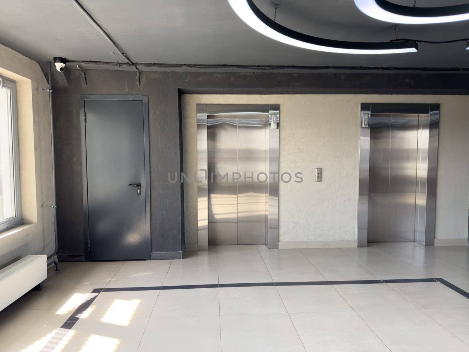 Modern building interior with two stainless steel elevators, dark utility door, surveillance camera and geometric ceiling light, illustrating clean minimalist hallway in residential or office space