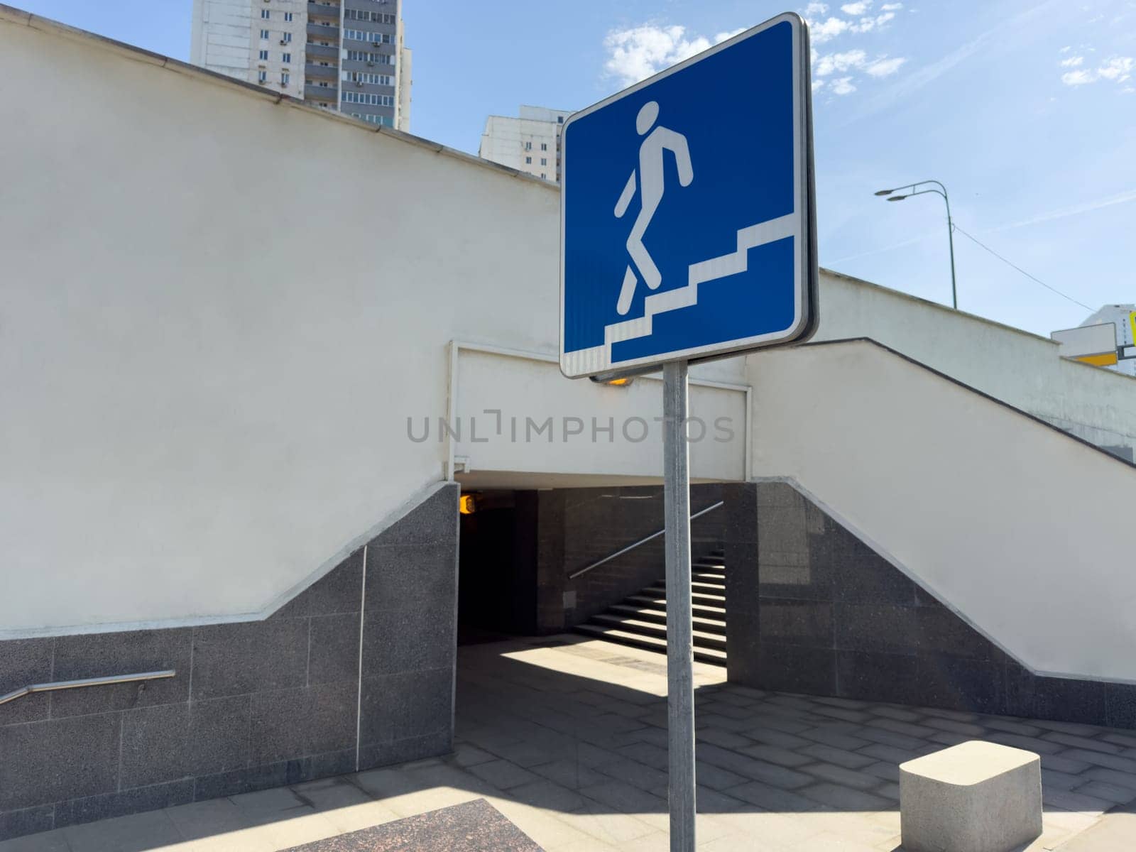 Blue pedestrian sign showing figure descending stairs placed in front of underpass entrance with white wall and tile base, representing urban navigation and public infrastructure in city environment