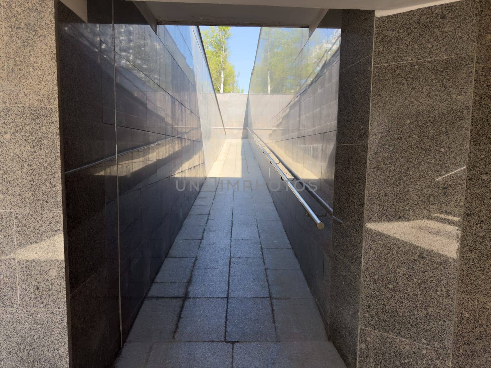 Brightly lit underground ramp with smooth stone floor and reflective granite walls leading to open sky, creating strong linear perspective and clean architectural composition in modern city passage