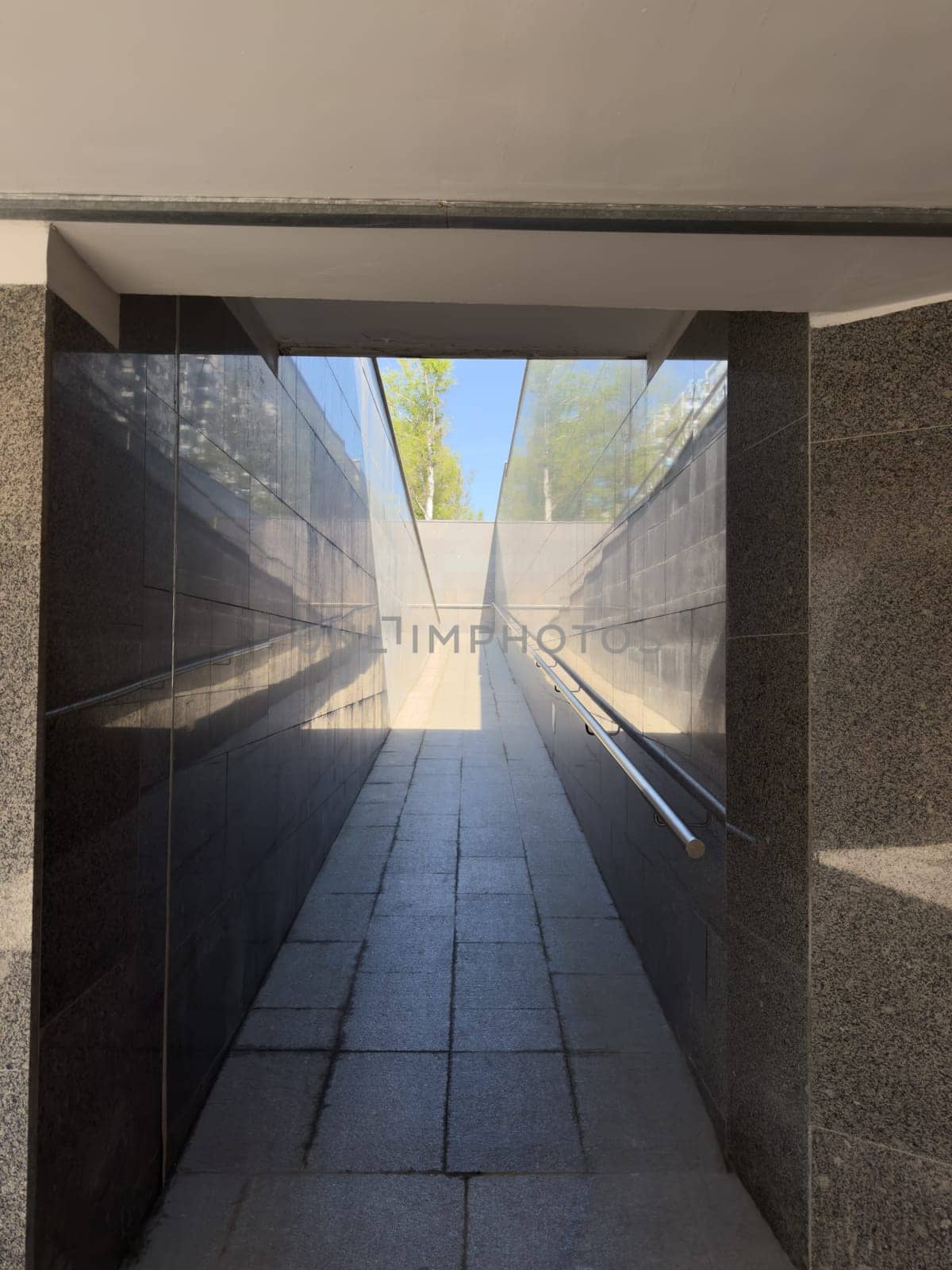 Gently inclined pedestrian ramp with smooth granite surface and handrails on both sides provides accessible pathway for wheelchairs and strollers in modern public infrastructure setting