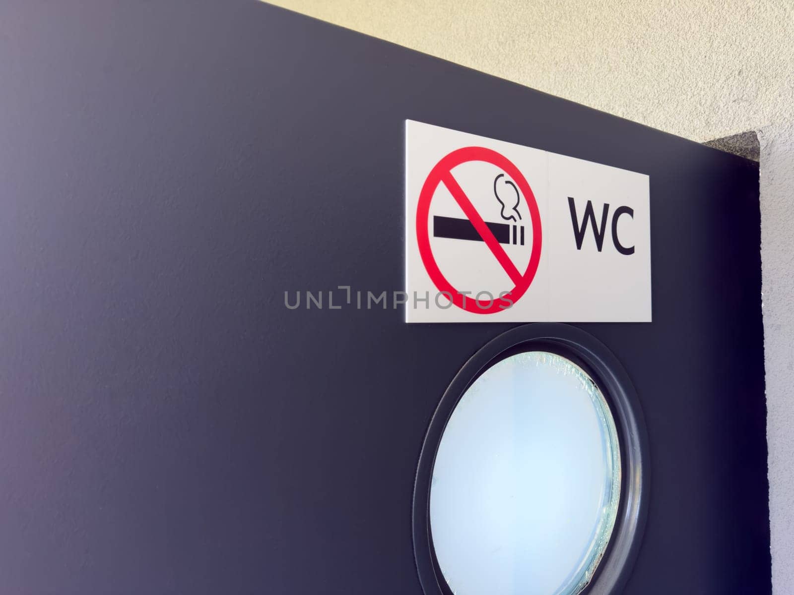 Closeup of WC door with round frosted window and no smoking sign on white label, showing clear visual message for public restroom users in clean and modern architectural setting