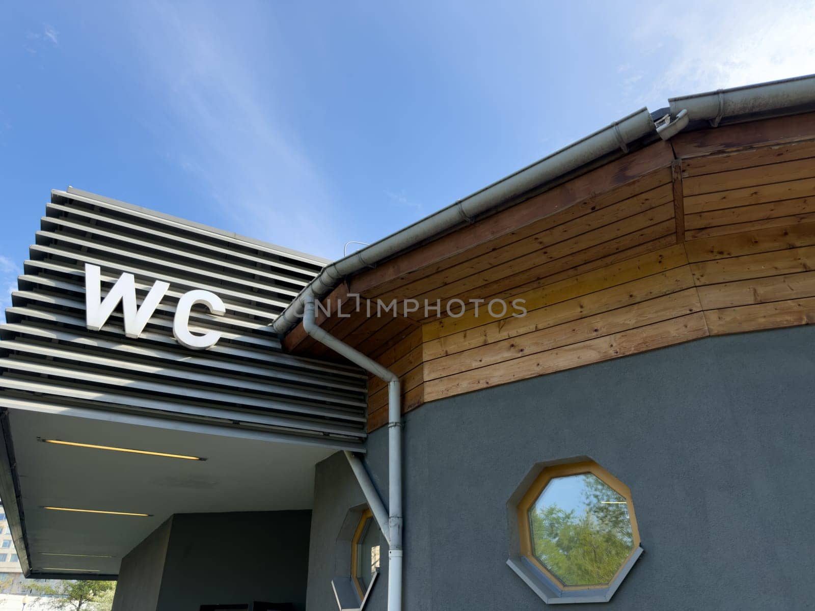 Modern public restroom building with large WC letters on metal facade and wooden roof detail under blue sky, combining practical function with clean architectural design in outdoor urban space