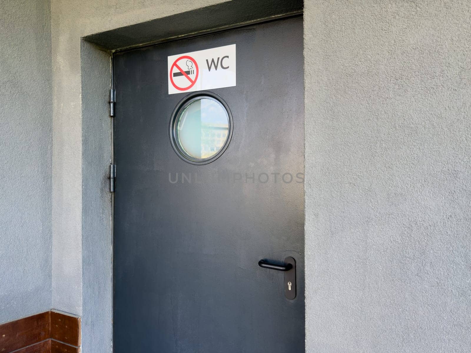 Black door with round frosted window and no smoking WC sign mounted on rough gray wall, marking entrance to public restroom with emphasis on clarity, hygiene and behavioral regulation in urban setting