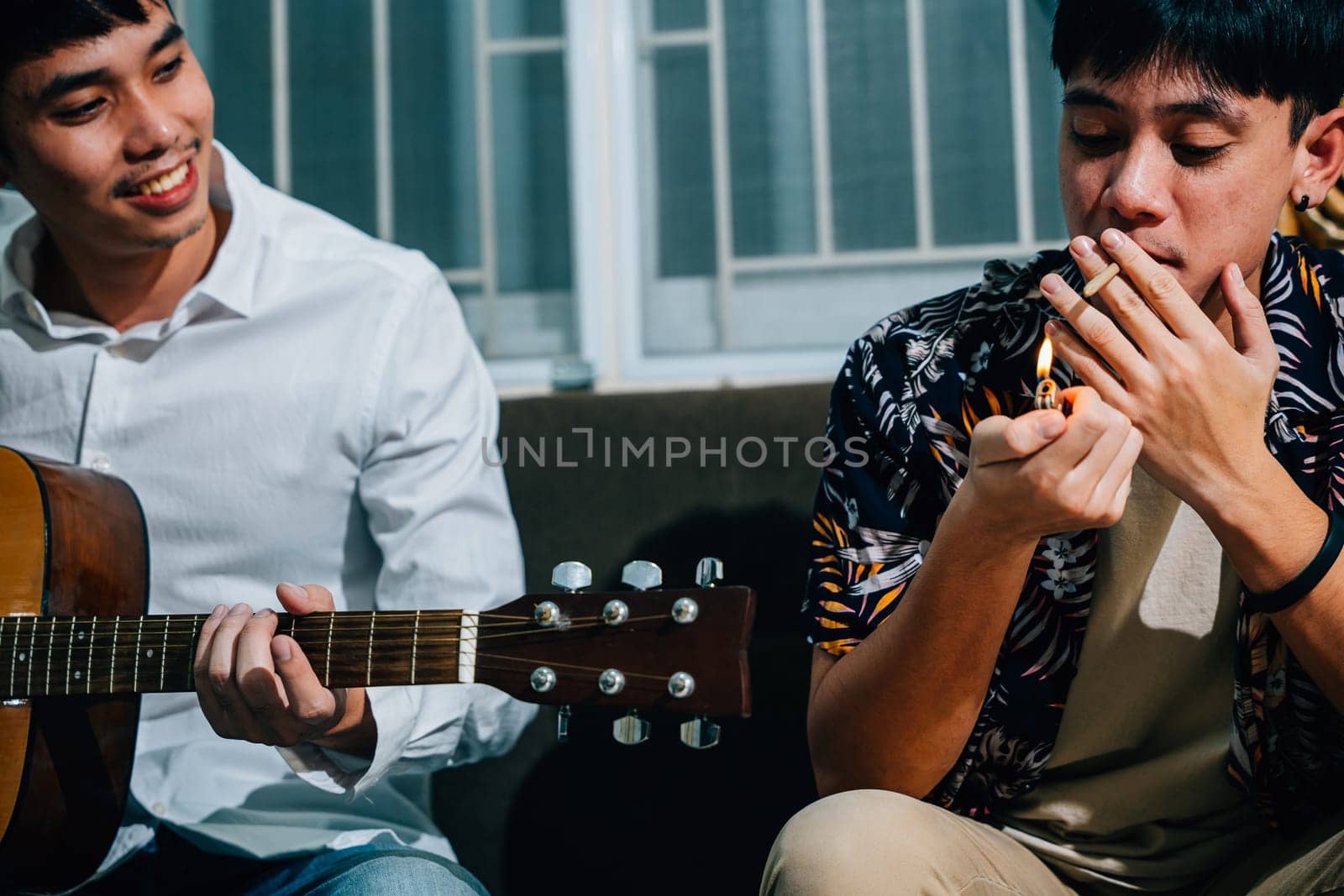 Two Asian men are sitting on a couch, one playing a guitar and the other smoking a cigarette while sitting on sofa in relaxing time at home. smoking marijuana or cannabis