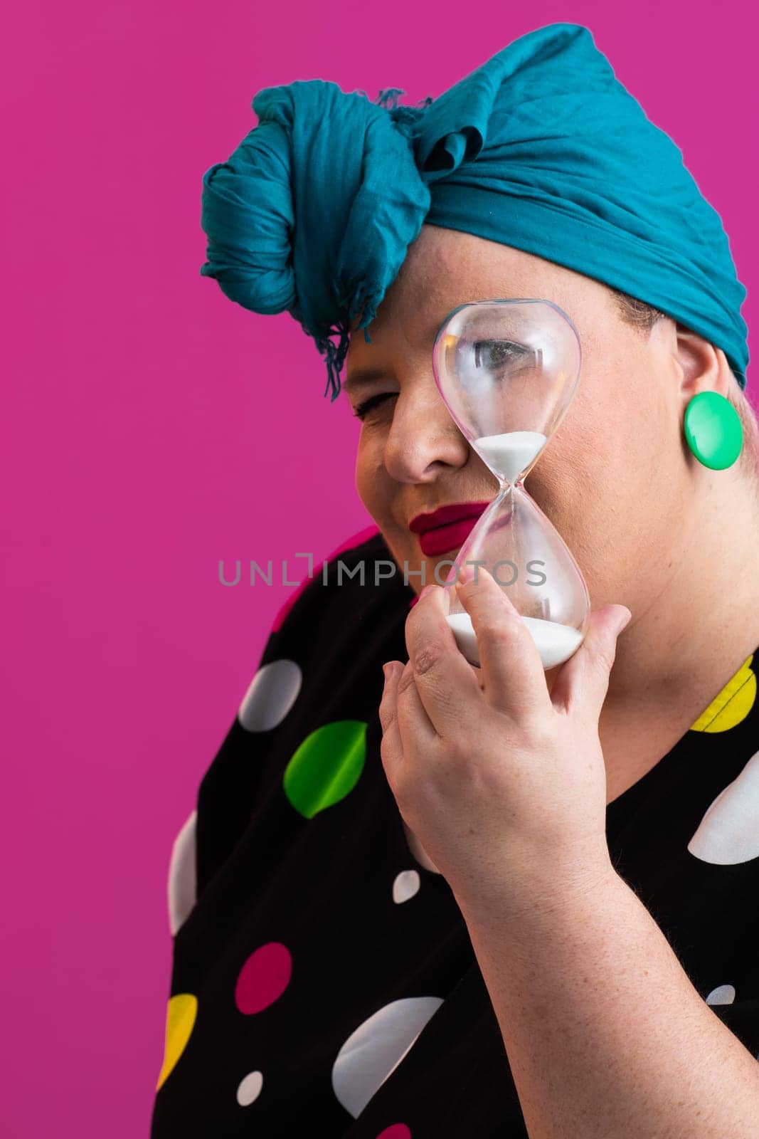 Portrait of a confident woman with a retro headwrap and colorful outfit, holding an hourglass in front of her face as a symbol of time and focus, with a vibrant pink background.