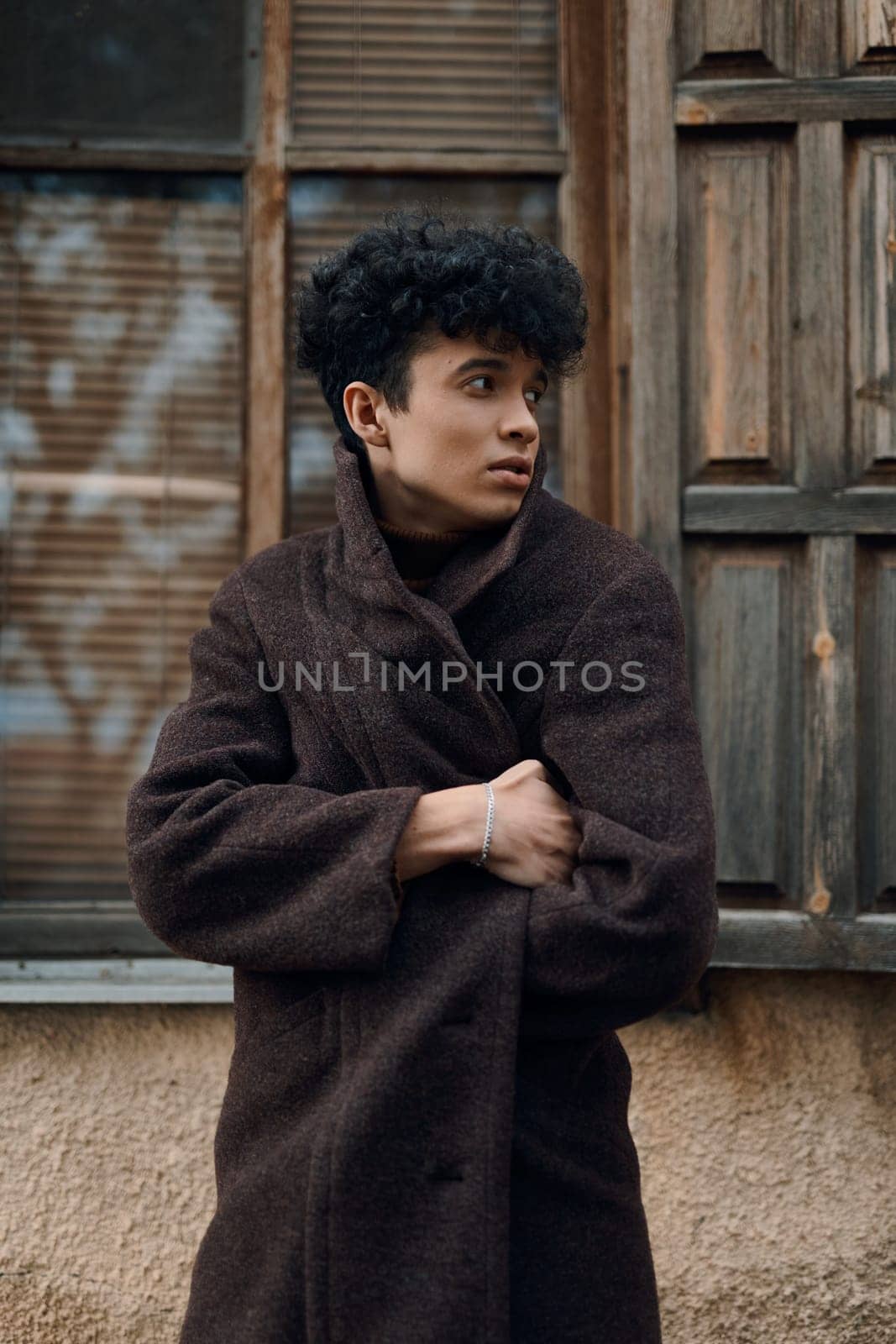 Fashionable young man with curly hair wearing a brown coat, standing in front of an old building with a window.