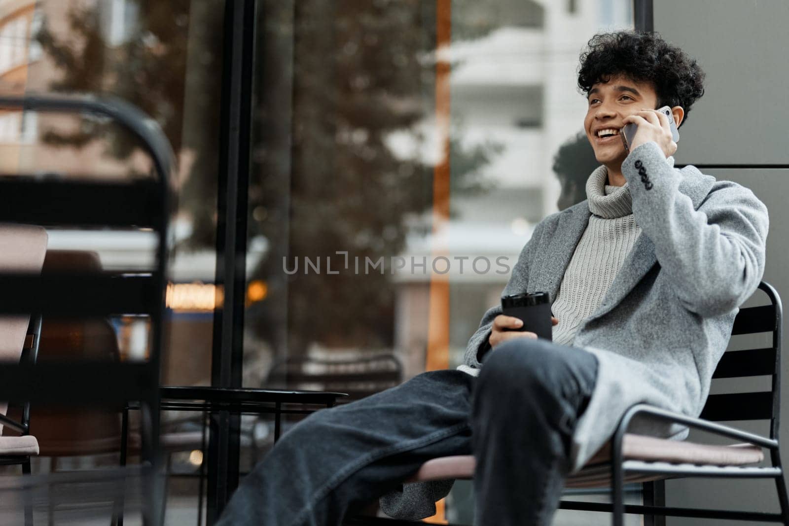 Man sitting on chair by glass window, talking on cell phone with coffee cup in hand. fashionable and stylish concept.