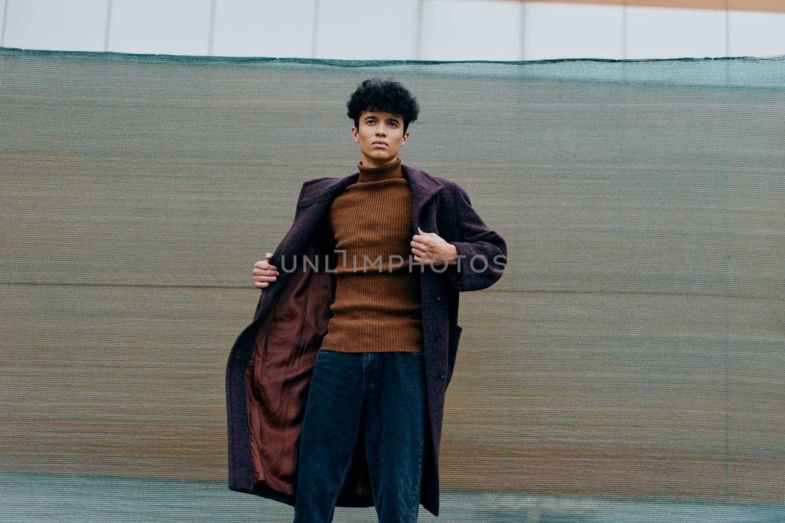 Fashion portrait of a stylish young man in a long coat and turtleneck sweater standing confidently with his hands in his pockets in front of a wall.