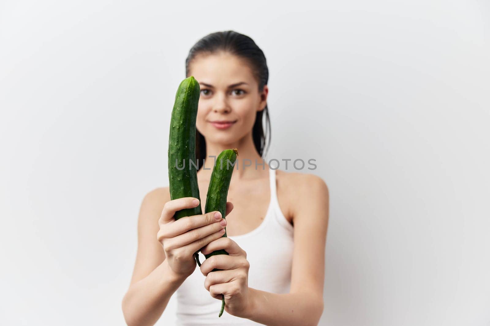 A young woman in a white tank top is cheerfully holding two fresh green cucumbers against a bright white background This image emphasizes health, vitality, and the importance of fresh produce in a healthy diet