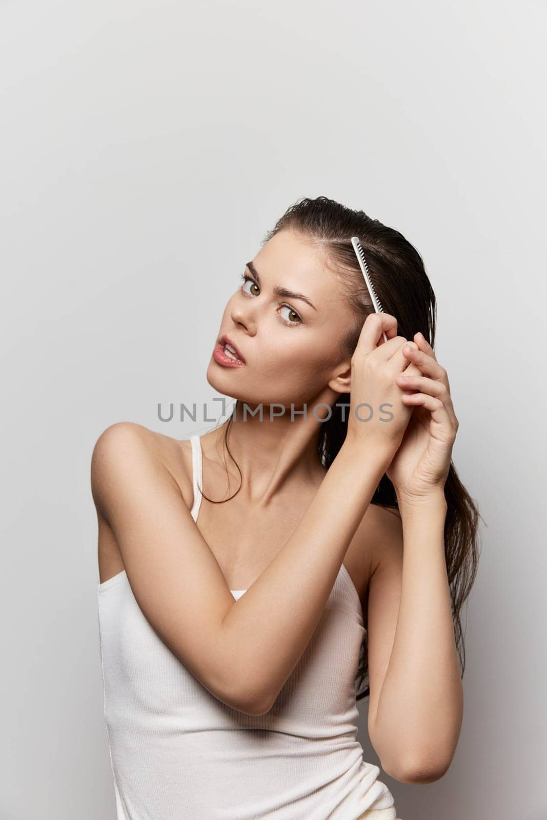 A young woman with wet hair is combing her hair with a fine toothed comb, showcasing a natural beauty routine She has a contemplative expression and is dressed casually against a light background