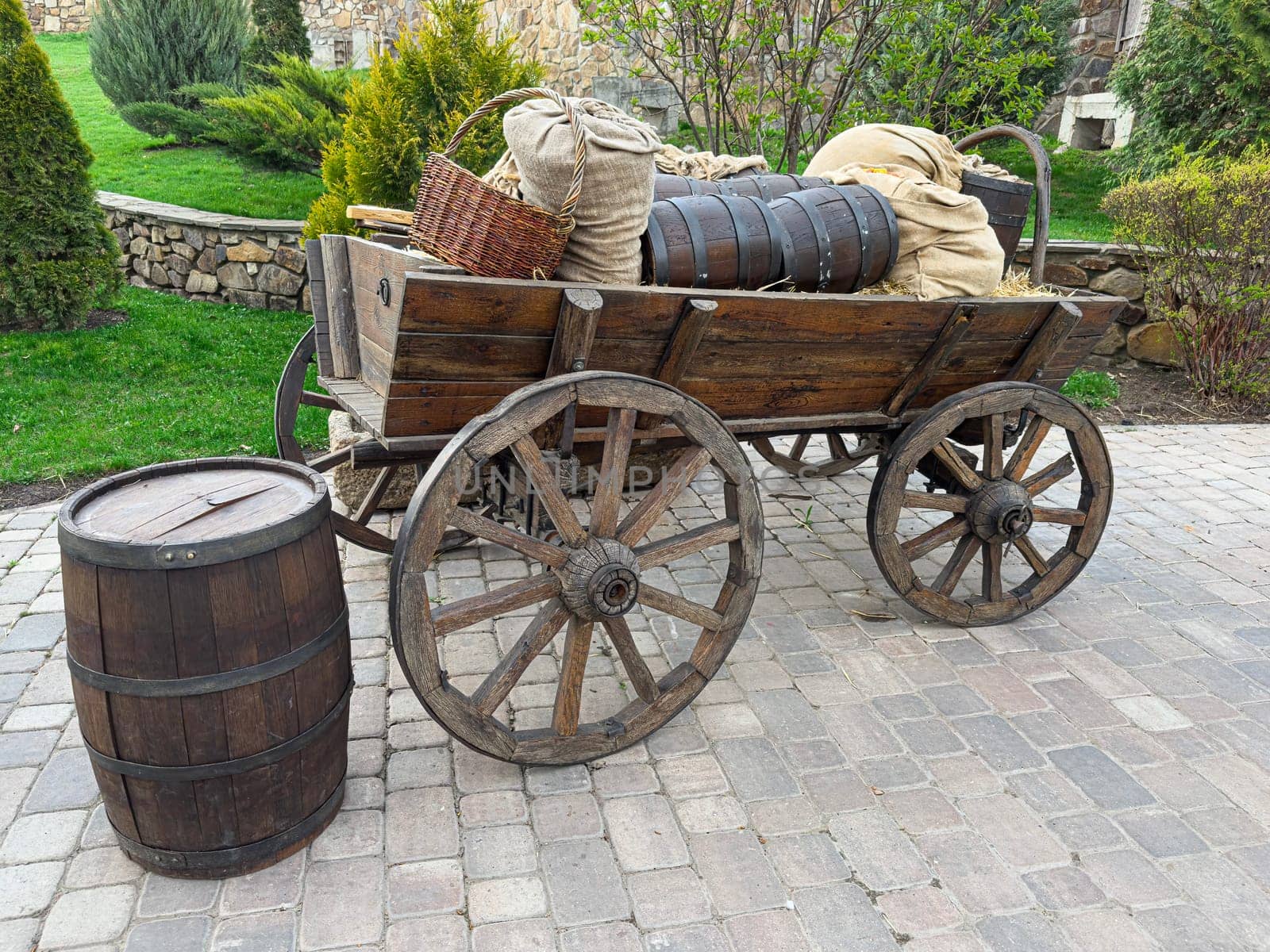 A rustic wooden cart stands in a garden, filled with barrels and burlap sacks, surrounded by lush greenery and stone landscaping. This scene evokes a nostalgic farming atmosphere.