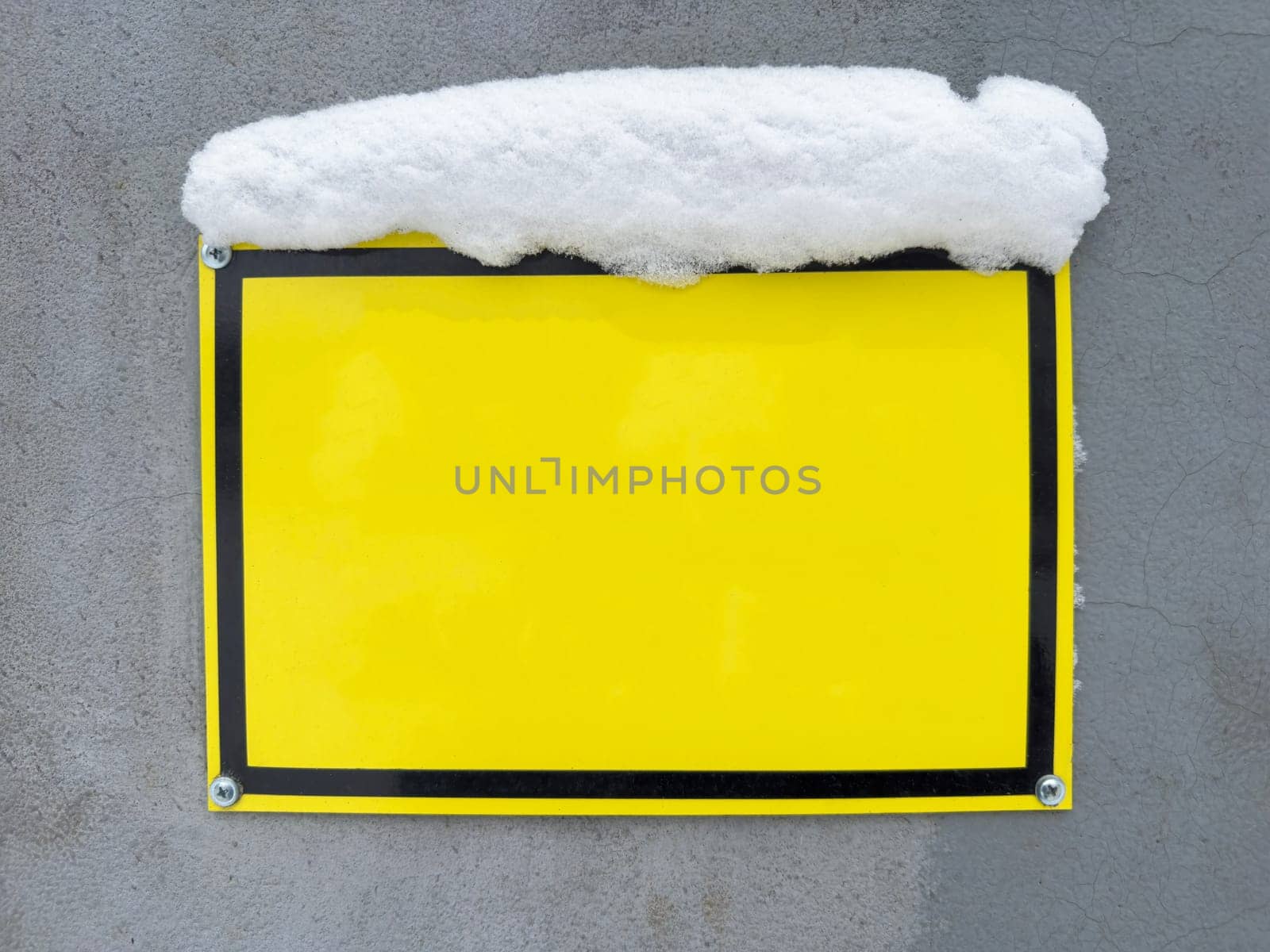 A vibrant yellow sign is attached to a gray wall, partially hidden under a layer of snow. The contrasting colors create a striking visual effect in a winter environment.
