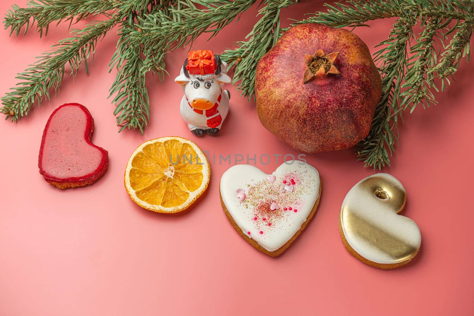 Colorful cookies shaped like hearts, dried orange slices, and a pomegranate are arranged with a small cow figurine and pine branches on a pink background, creating a joyful holiday display.