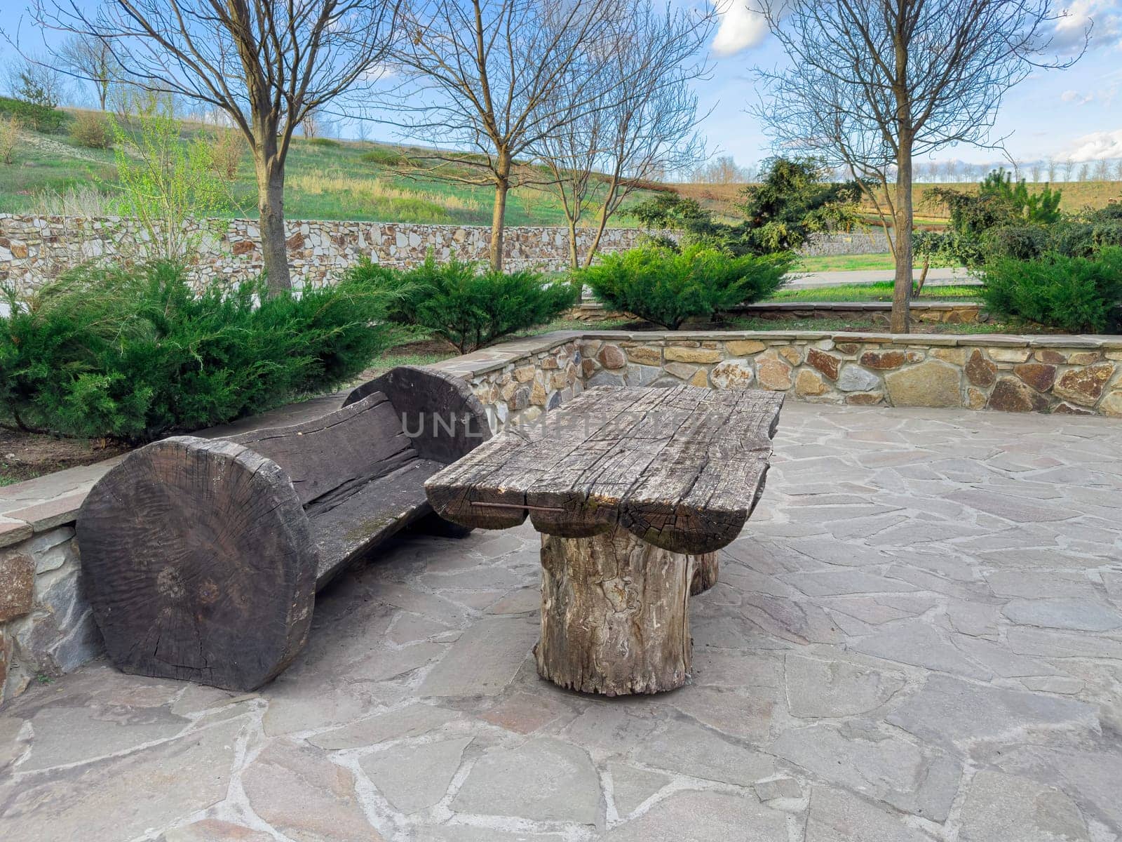 Wooden bench and table made from natural materials sit on a stone patio in a quiet garden. Lush greenery surrounds the area under a clear blue sky, creating a peaceful atmosphere.