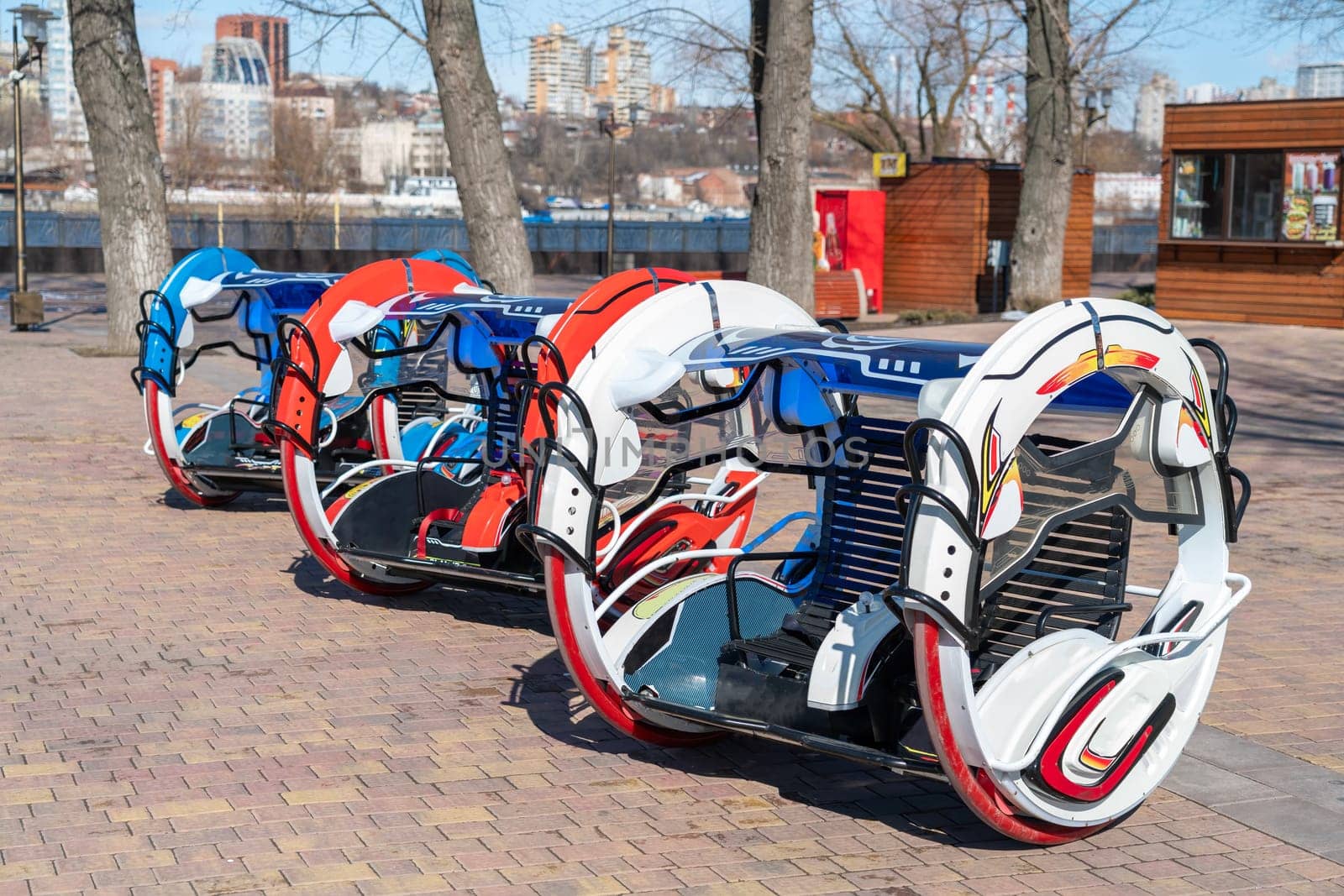 Colorful hover-like vehicles line the riverside walkway in a city, showcasing modern design. The area is sunny with trees and modern buildings in the background, indicating a vibrant setting.