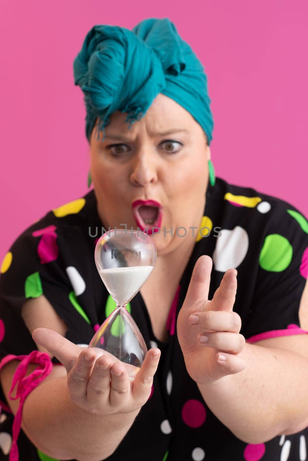 Portrait of a confident woman with a retro headwrap and colorful outfit, holding an hourglass in front of her face as a symbol of time and focus, with a vibrant pink background.
