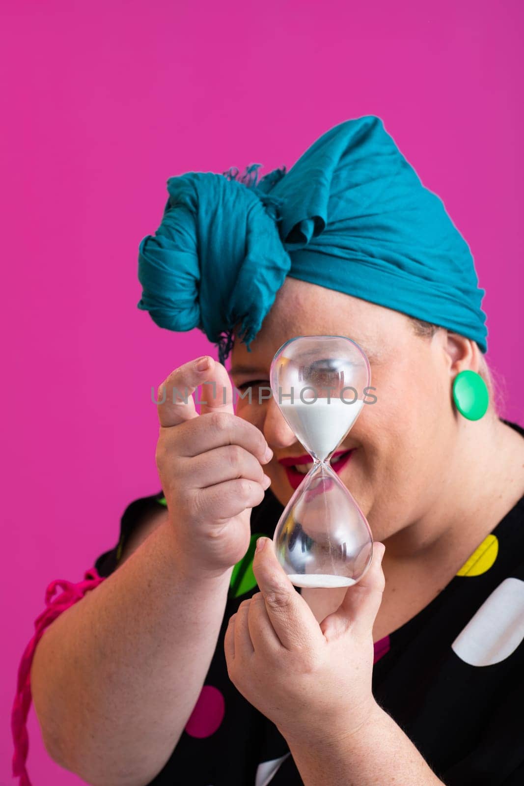 Portrait of a confident woman with a retro headwrap and colorful outfit, holding an hourglass in front of her face as a symbol of time and focus, with a vibrant pink background.