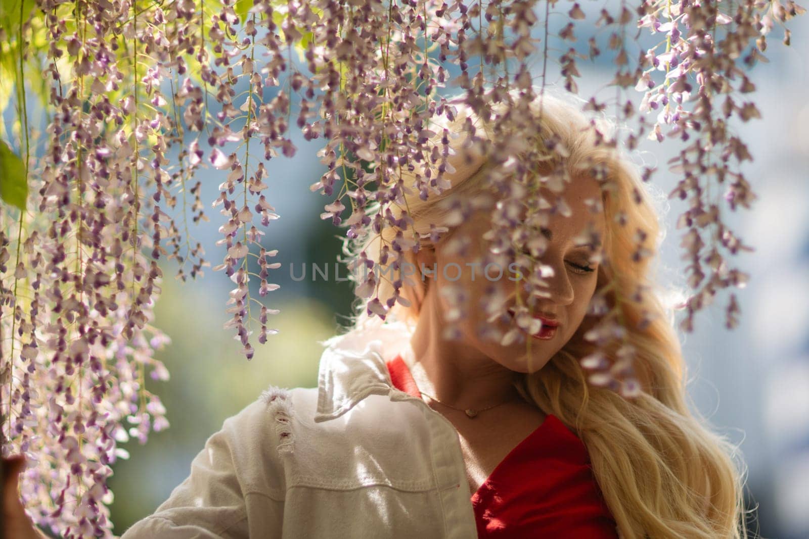 Blonde woman in red top stands under cascading wisteria. Natural light, soft focus. Relaxed pose in a garden setting.