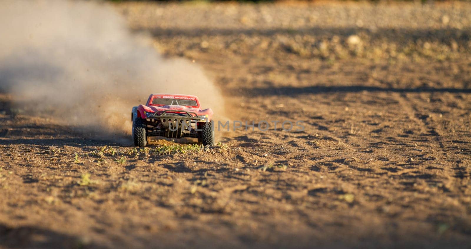 A red toy truck is driving through a dusty field. The truck is covered in dirt and the ground is covered in dust