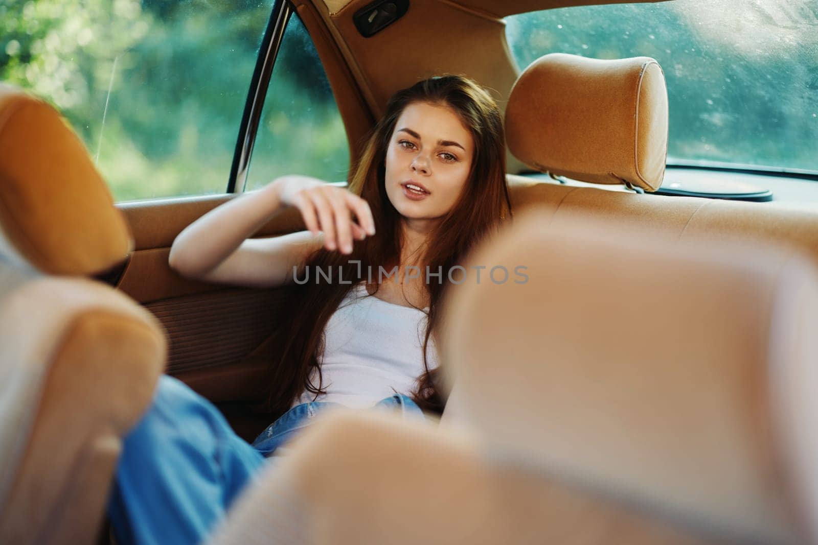 A young woman with long hair is lounging in the back seat of a vintage car, conveying a relaxed and carefree vibe amidst a beautiful green backdrop, perfect for moods of summer and freedom.