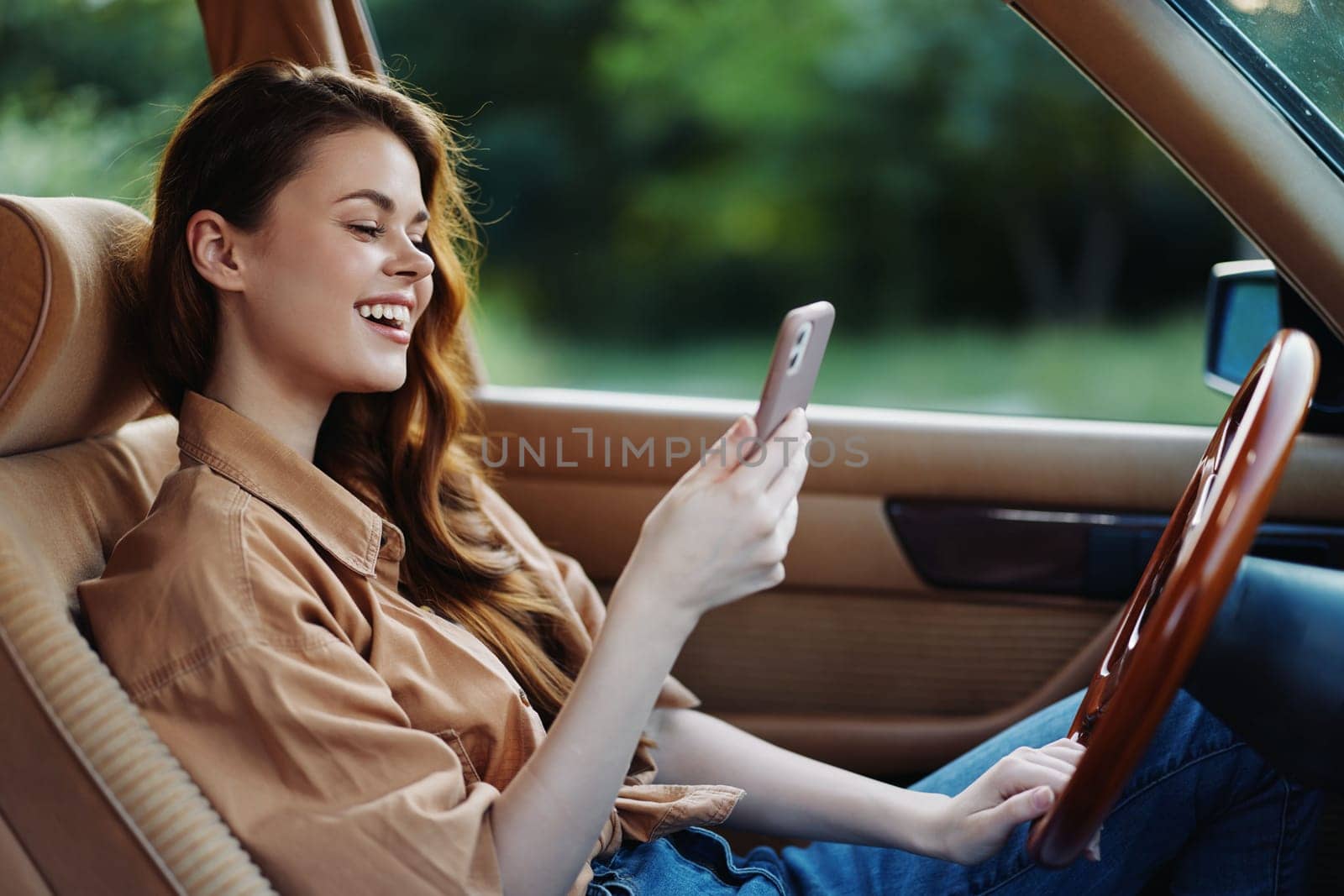 A joyful young woman in a casual brown shirt smiling while using her smartphone in a vintage car with a lush green background, reflecting happiness and leisure.