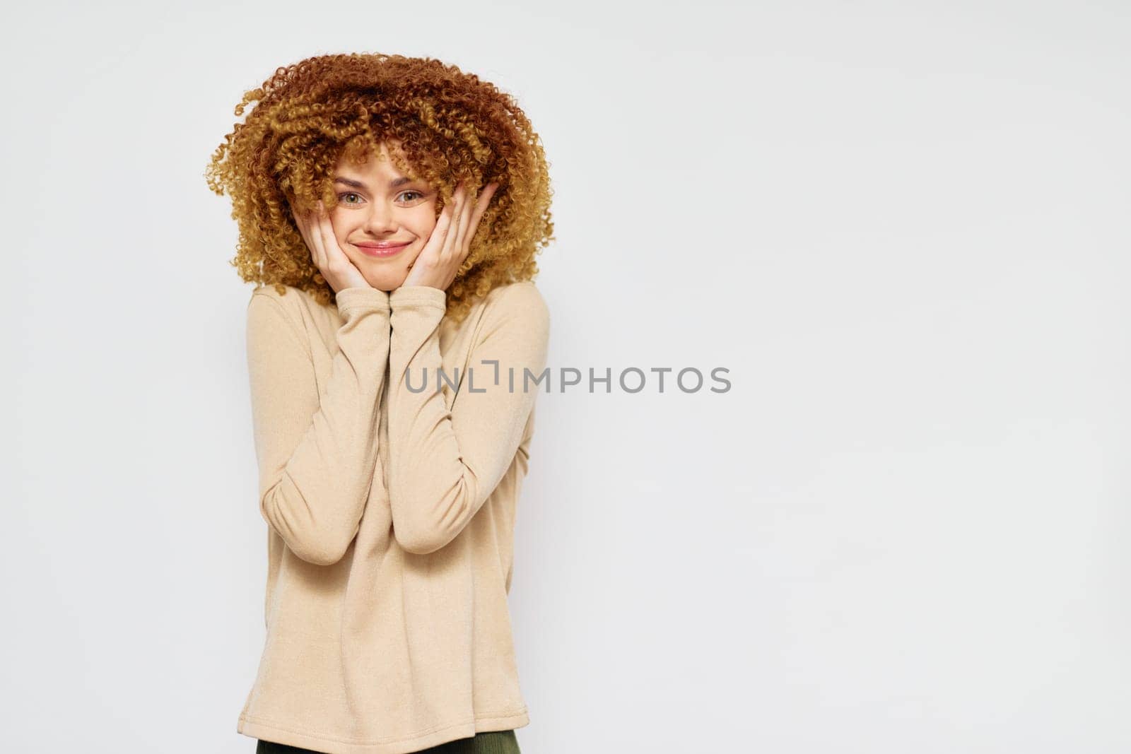 Happy young woman with curly hair in a beige shirt, displaying a cheerful demeanor She stands against a minimalist white backdrop, conveying a sense of joy and positivity through her playful express