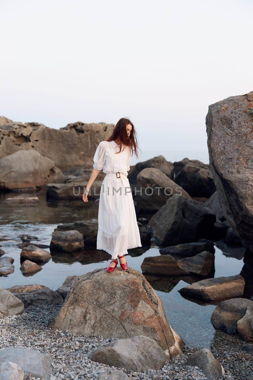Serene white dress woman with red shoes standing on rock near water's edge