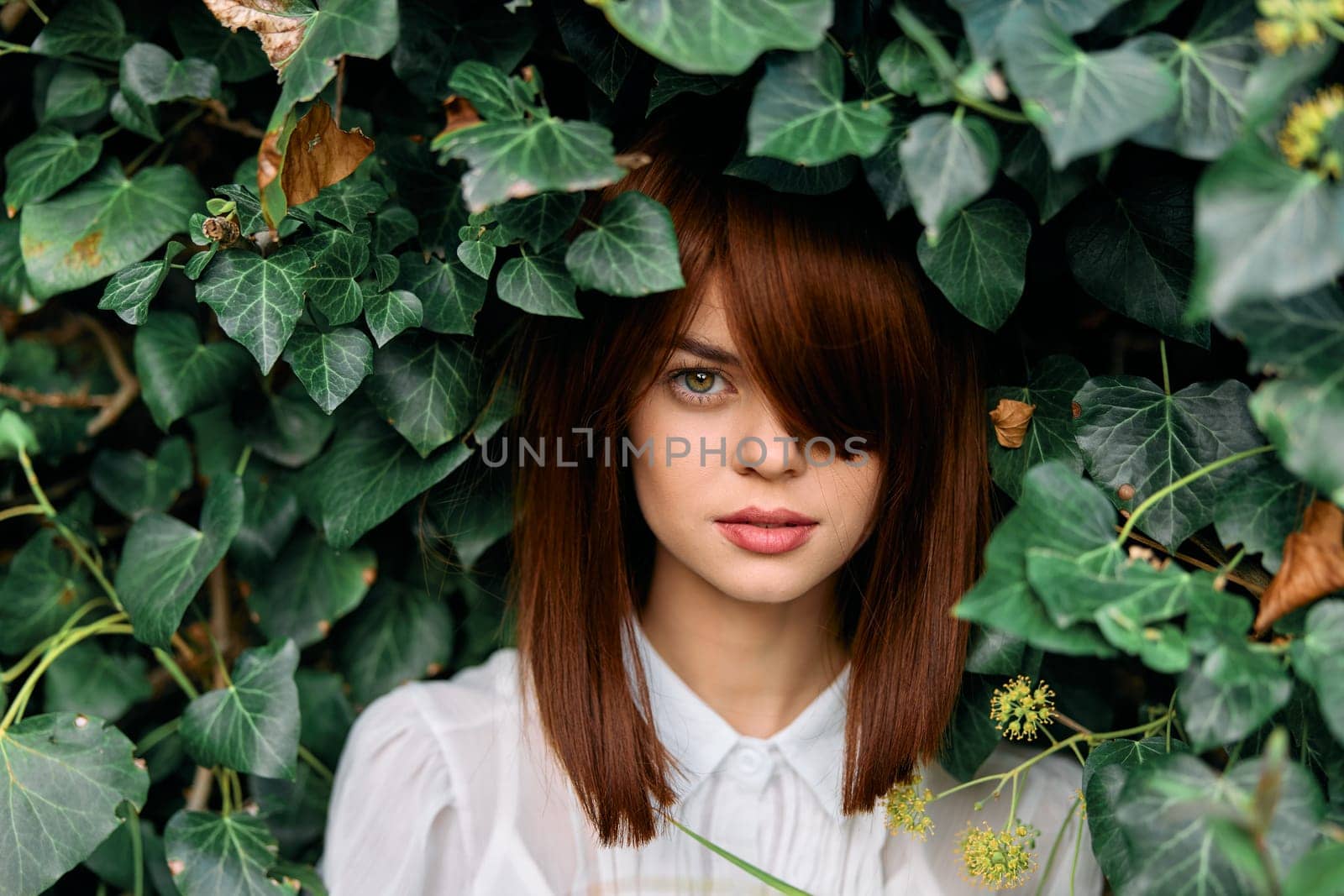 Redhaired woman in white shirt surrounded by lush green ivy foliage in outdoor setting