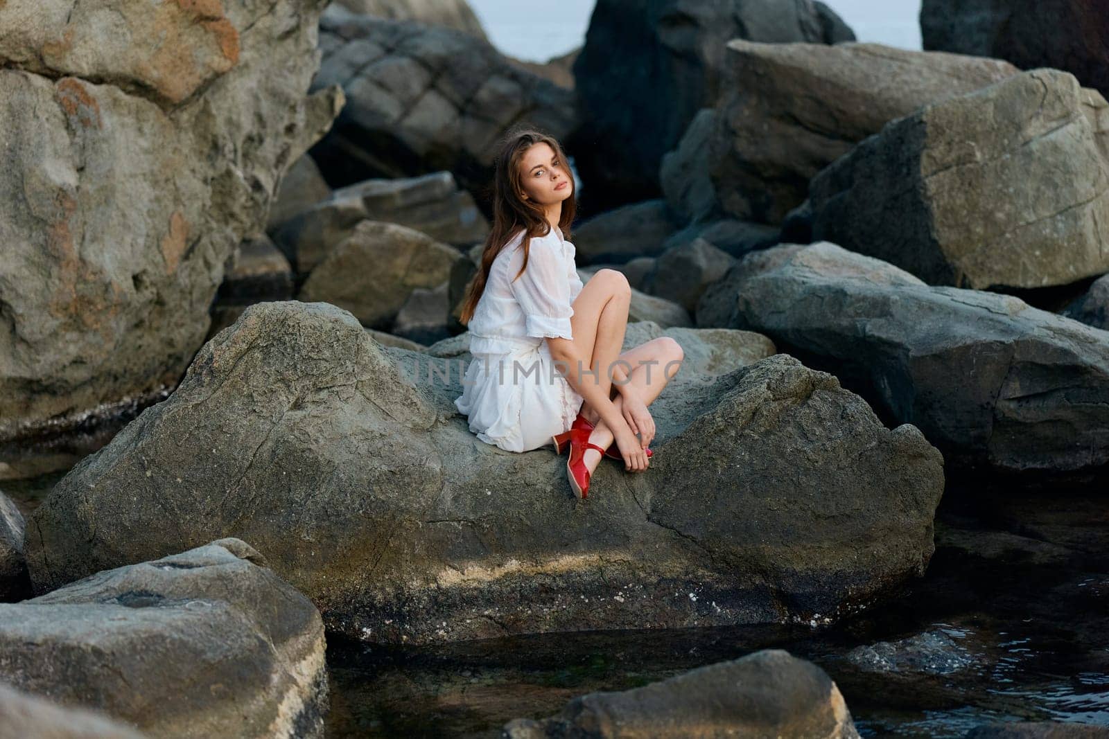 Serene woman in white dress relaxing on coastal rock with bright red shoes alongside ocean waves