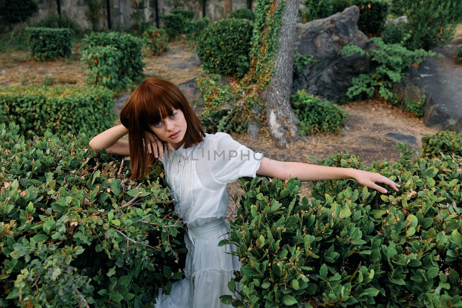 Serene young woman in white dress leaning against lush hedge in tranquil park setting