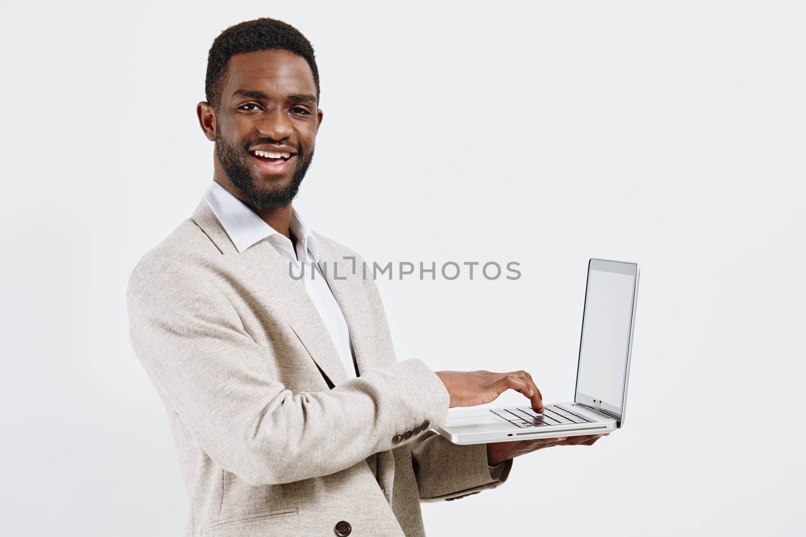 Cheerful man with dark skin, dressed in smart casual beige cardigan and white shirt, holding a silver laptop in hand, smiling confidently against plain white background, professional and friendly appearance. Concept: modern business success