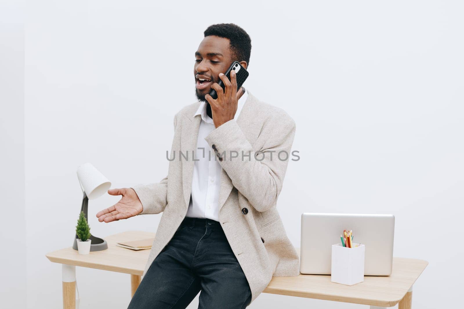 Happy man engaging in conversation on mobile phone in contemporary office with desk, laptop, and decorative elements, dressed casually yet professionally, expressing positive emotions and effective communication, workspace environment, lifestyle.