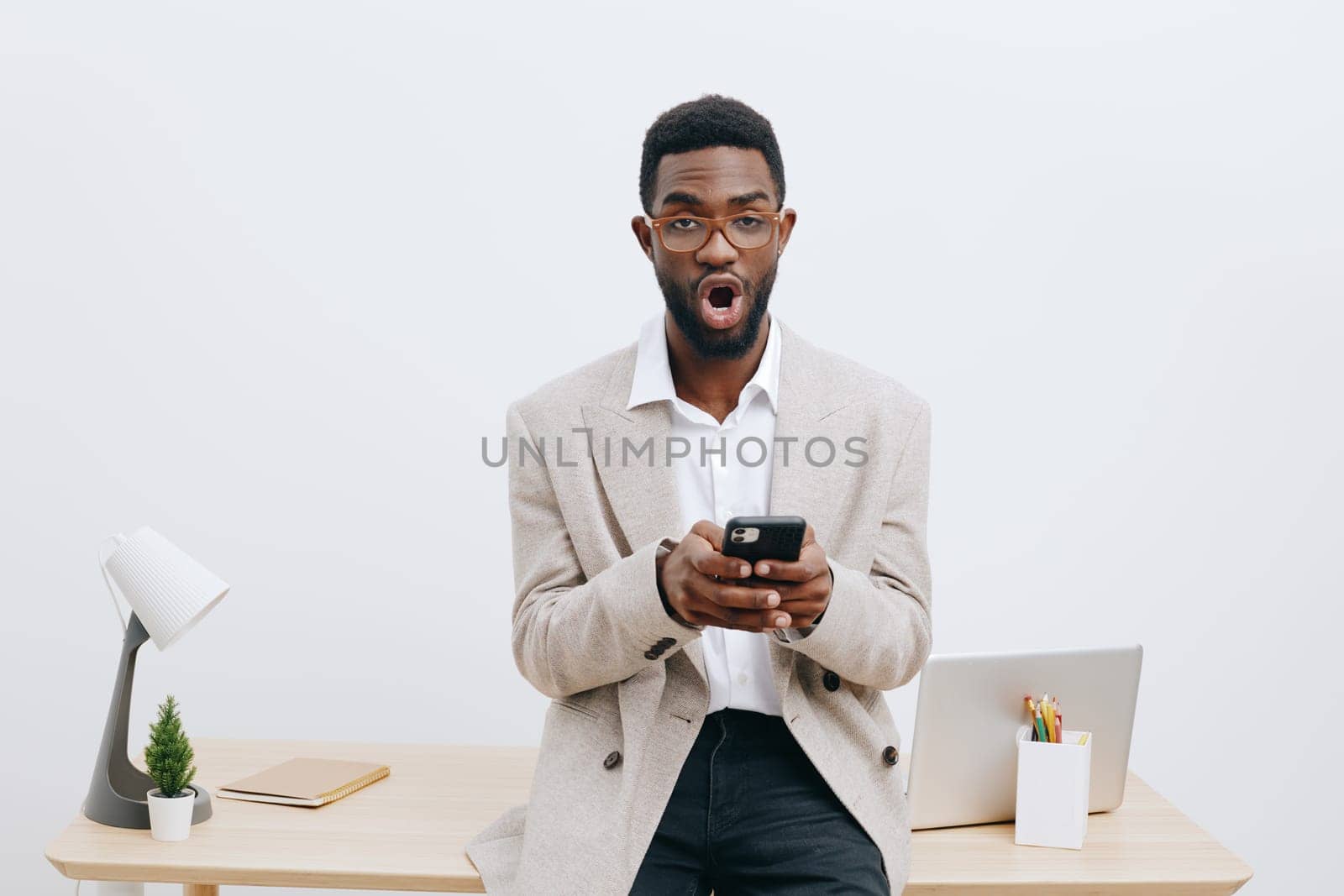 Happy man holding smartphone in bright modern office, surprised and excited mood, dressed casually in beige blazer and white shirt, office desk with laptop, stationery, minimal decor, clean white wall, workplace environment, lifestyle concept.
