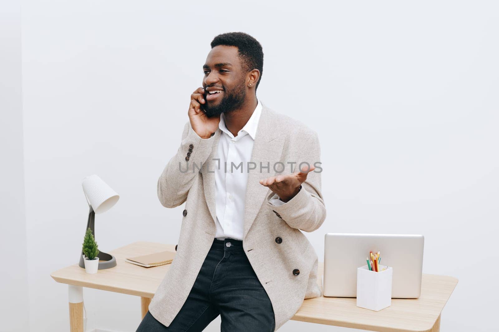 Happy young man with dark skin wearing beige blazer and white shirt, smiling and talking on mobile phone while sitting at a desk in a bright minimalistic office setting. Concept: communication and professional success.