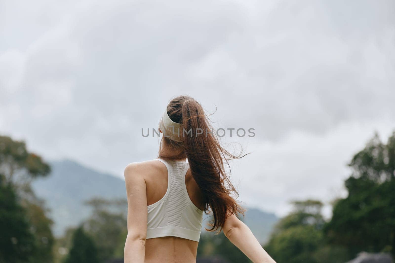 A fit woman from a Caucasian background wearing a sporty outfit stretches her arms joyfully outdoors The natural background and cloudy skies highlight health and vitality