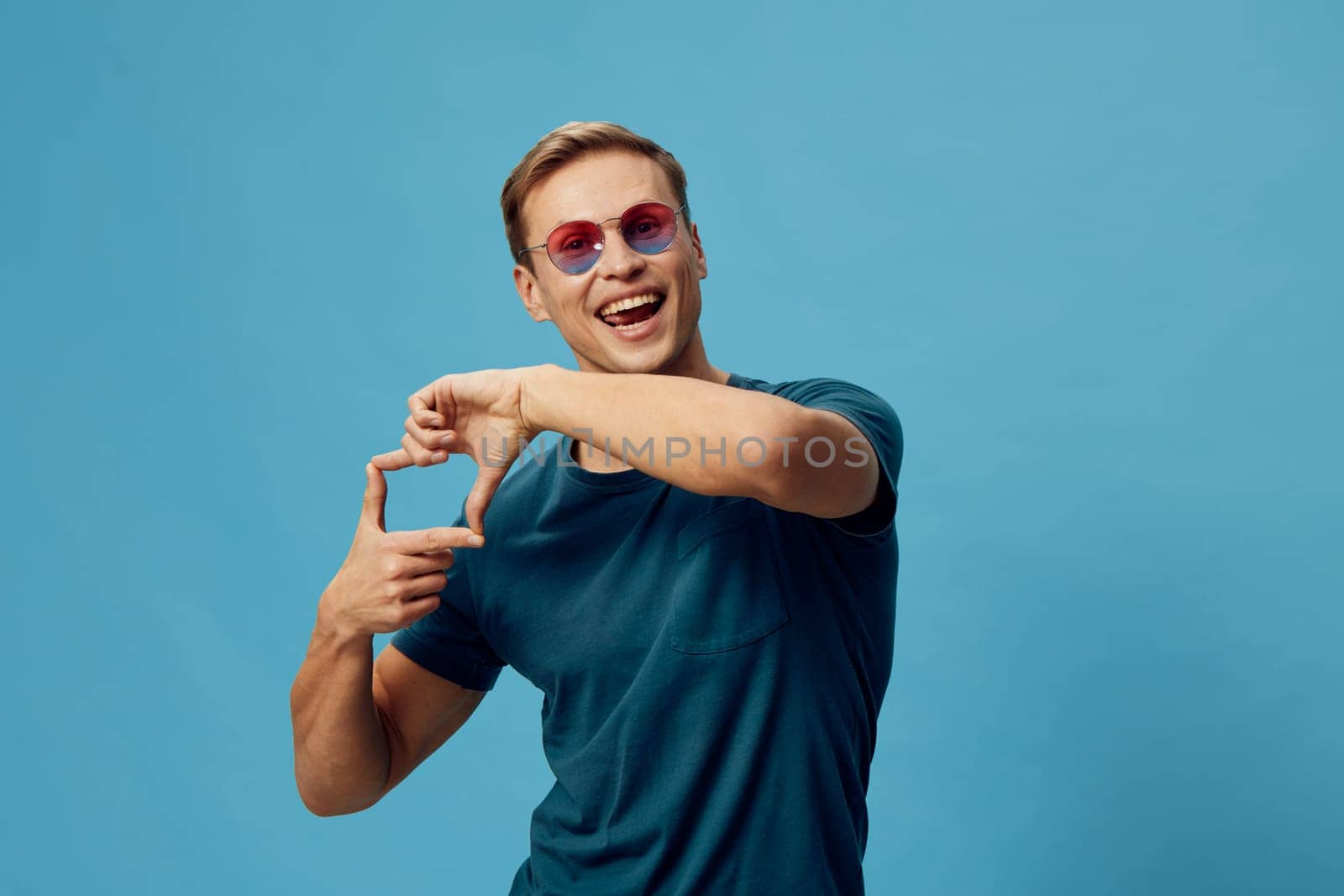 Smiling young caucasian man wearing blue t-shirt and pink sunglasses making frame gesture with hands isolated on plain blue background. Man expressing creativity and content generation using digital media. Concept: digital content creation.