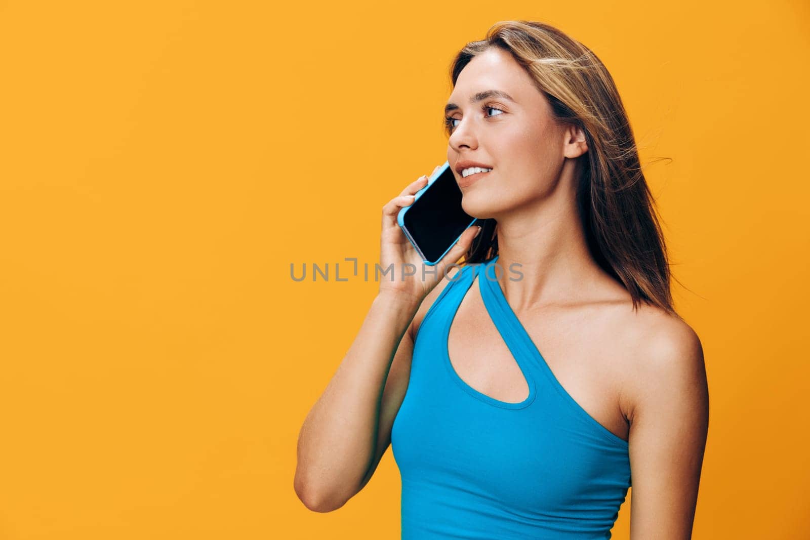 Vibrant woman in blue tank top engaged in a lively phone conversation against bright yellow backdrop
