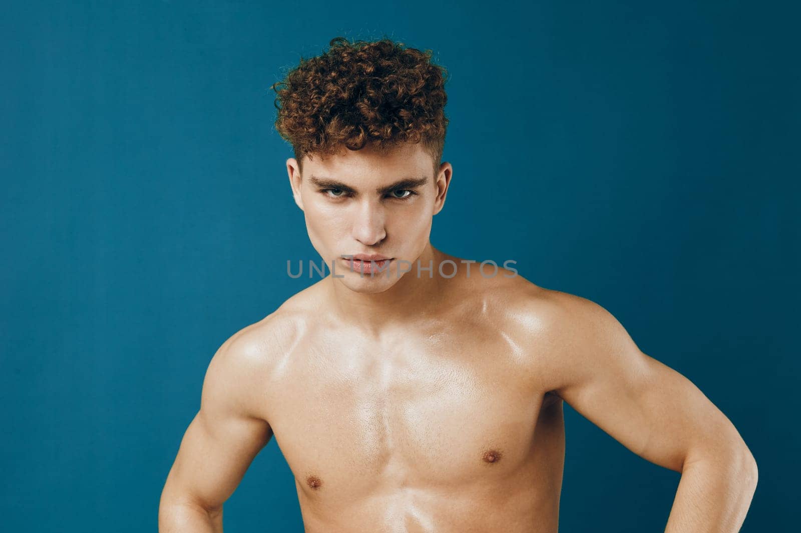 Strong male model with curly hair stands confidently topless against a vibrant teal background, embodying strength and self-assurance.