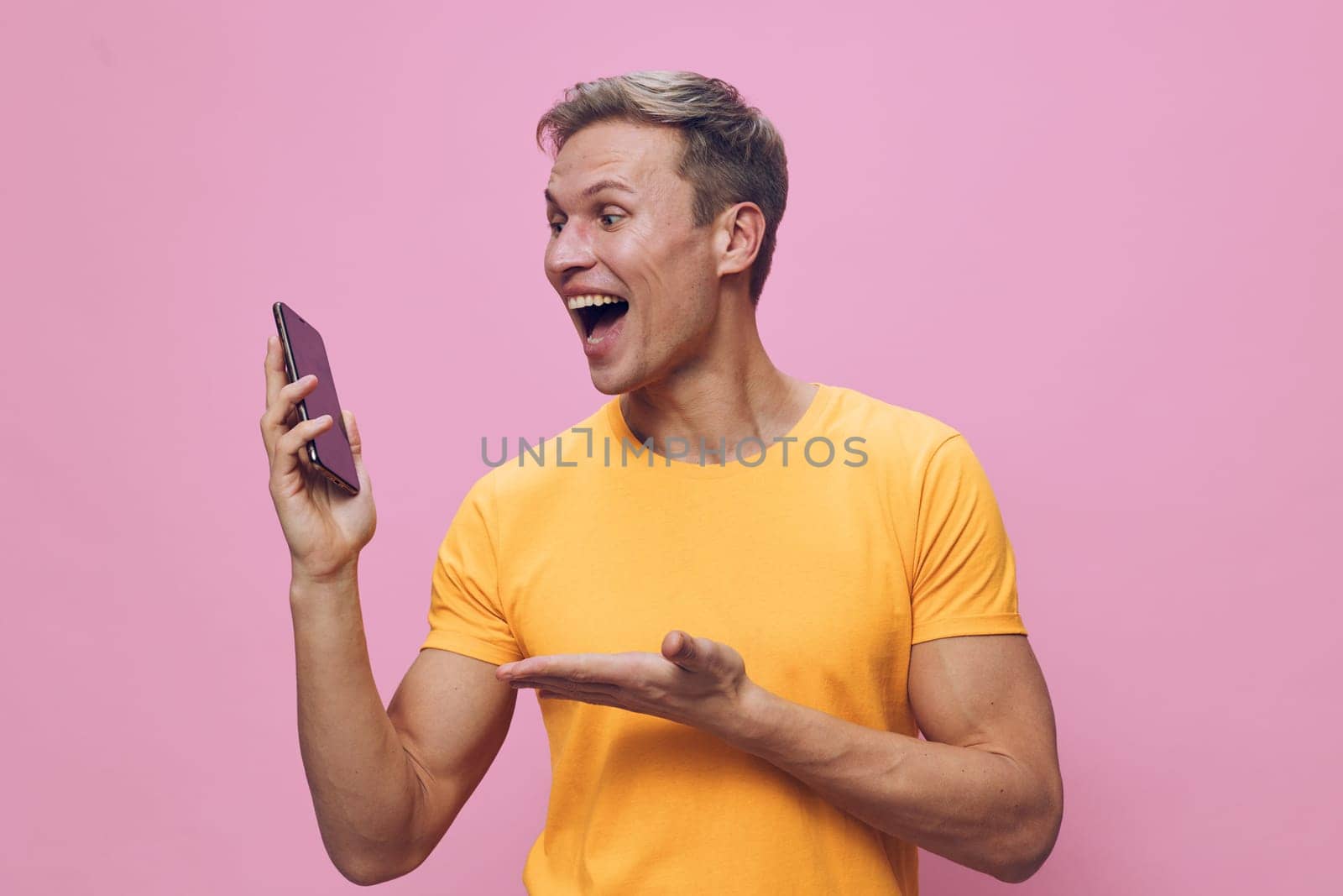 Cheerful man with short hair in yellow T-shirt looking at smartphone with happy expression isolated on pink background, showing surprise or excitement. Concept: social media, communication, technology