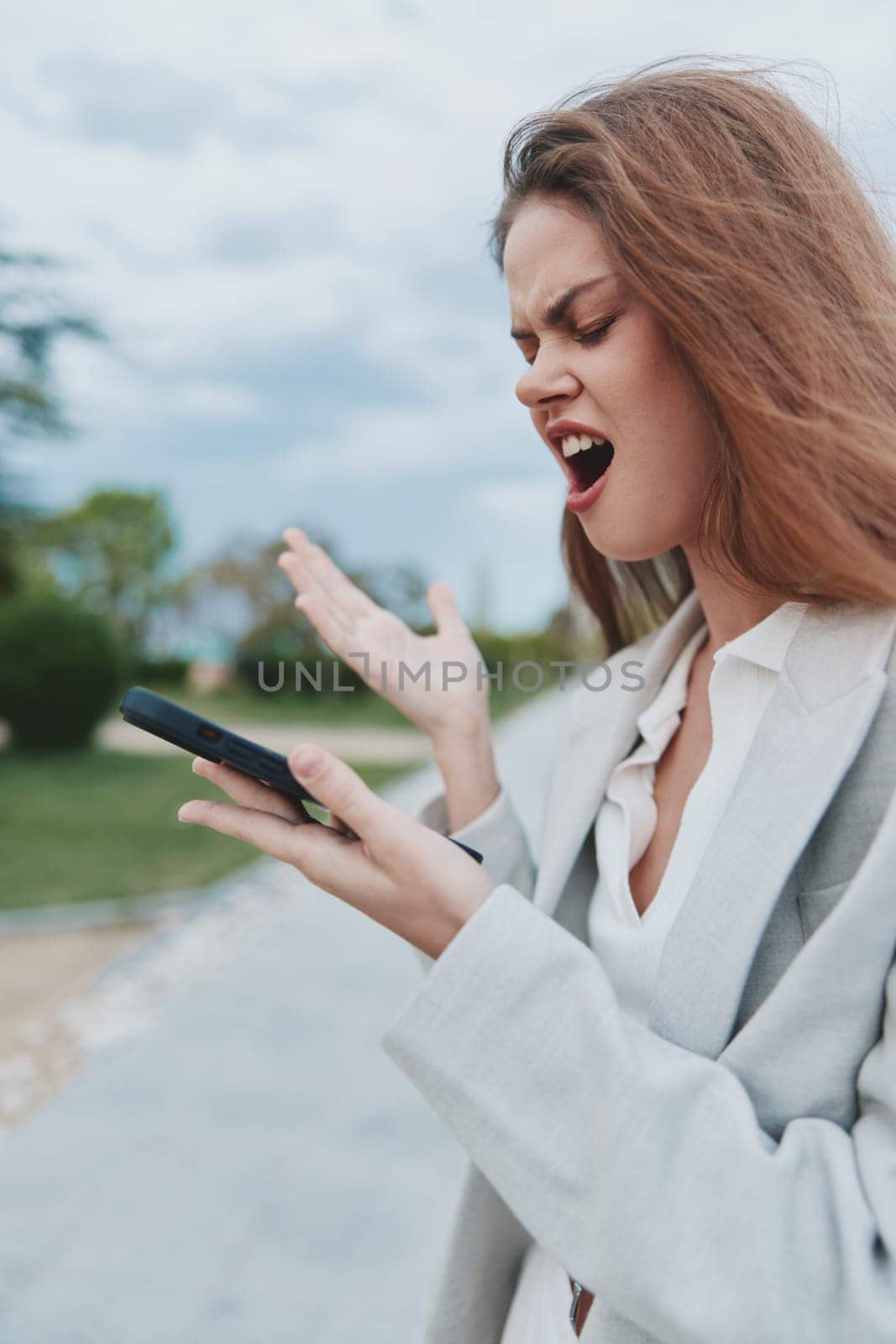 frustrated woman in casual business attire expressing anger and stress outdoors while looking at smartphone, symbolizing financial crisis and emotional struggle.