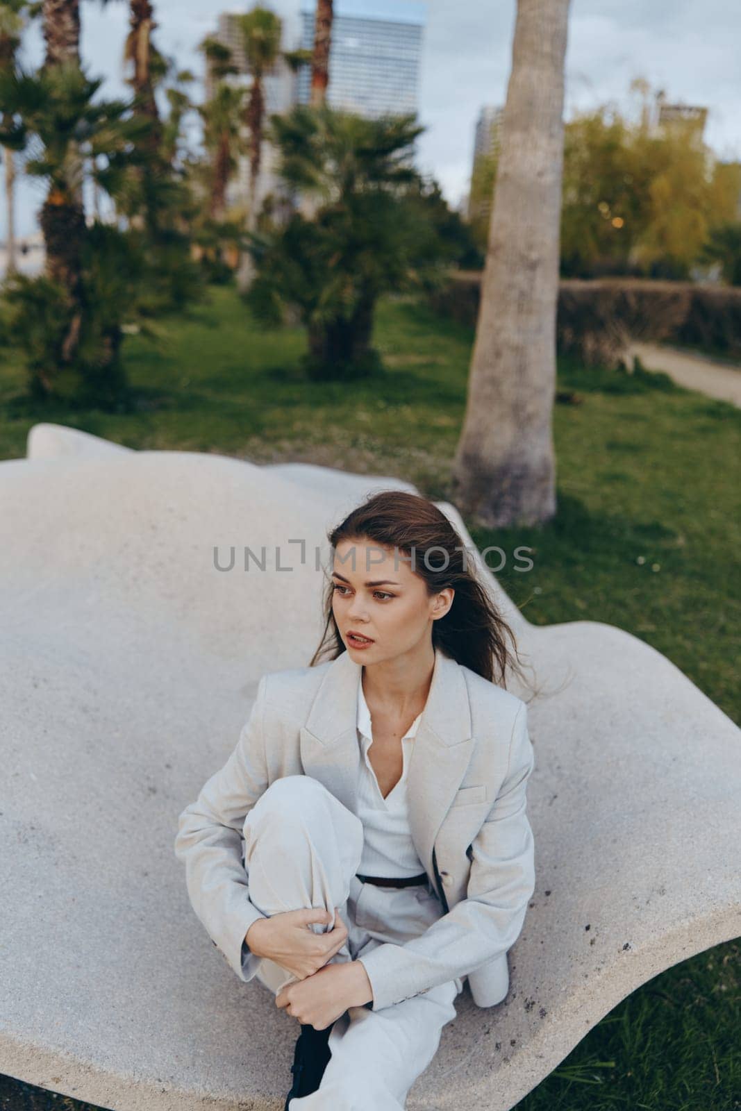 Young woman in light suit sits thoughtfully on curved stone bench outdoors in spring park, evoking fashion and psychological concept with natural green background.