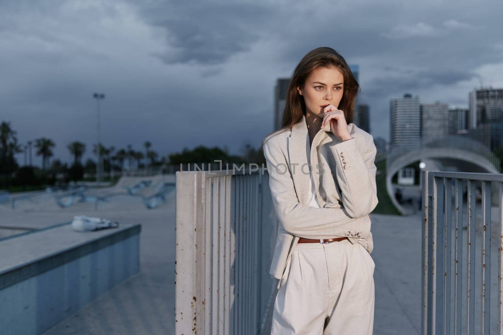 Thoughtful woman in beige business suit stands outdoors with modern cityscape and cloudy sky background, showing focus and contemplation in urban business setting