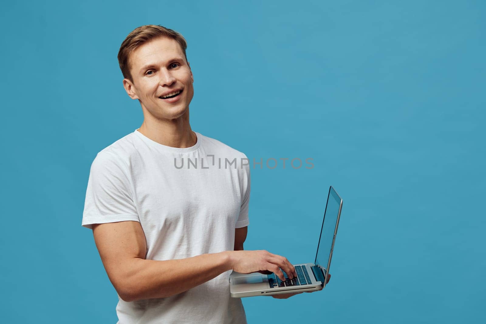 Happy young man in casual clothing holding a laptop computer against a plain blue background, smiling at camera, relaxed pose, technology and communication concept, studio portrait, modern lifestyle, digital device, cheerful expression, casual fashion.