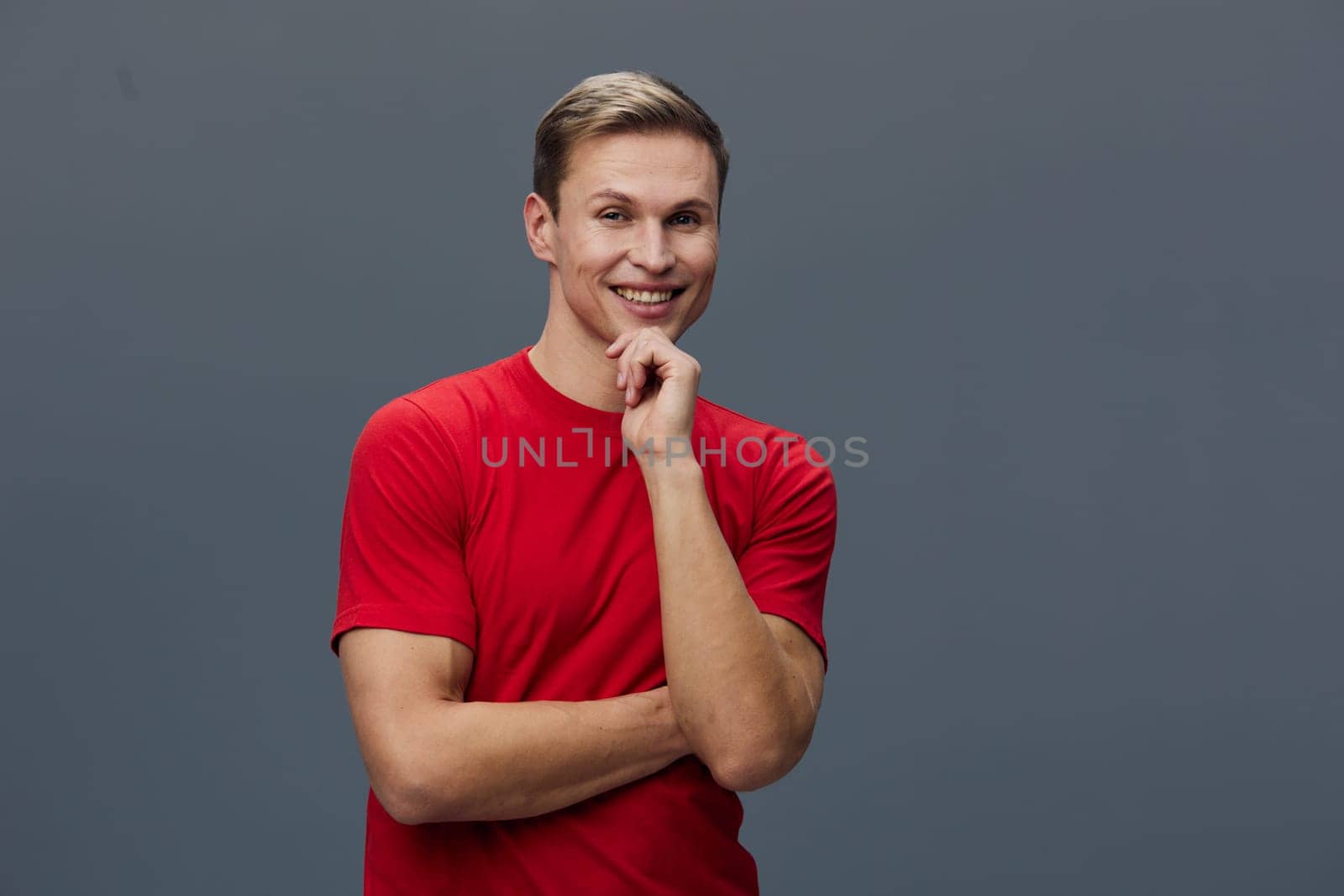 Portrait of a smiling young man wearing a red t-shirt, standing with arms crossed and hand on chin, expressing happiness and confidence, simple gray backdrop, casual style, cheerful mood.