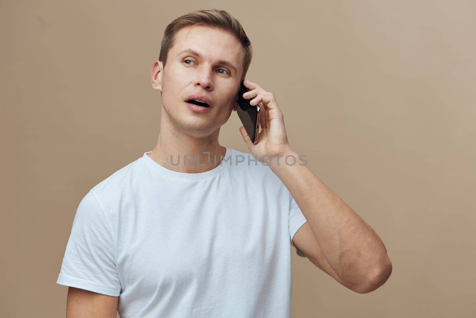 Close-up of a smiling young man with short hair, casually dressed in a light blue t-shirt, holding a mobile phone to his ear, reacting with astonishment as he talks, against a neutral beige backdrop. Concept: communication and surprise