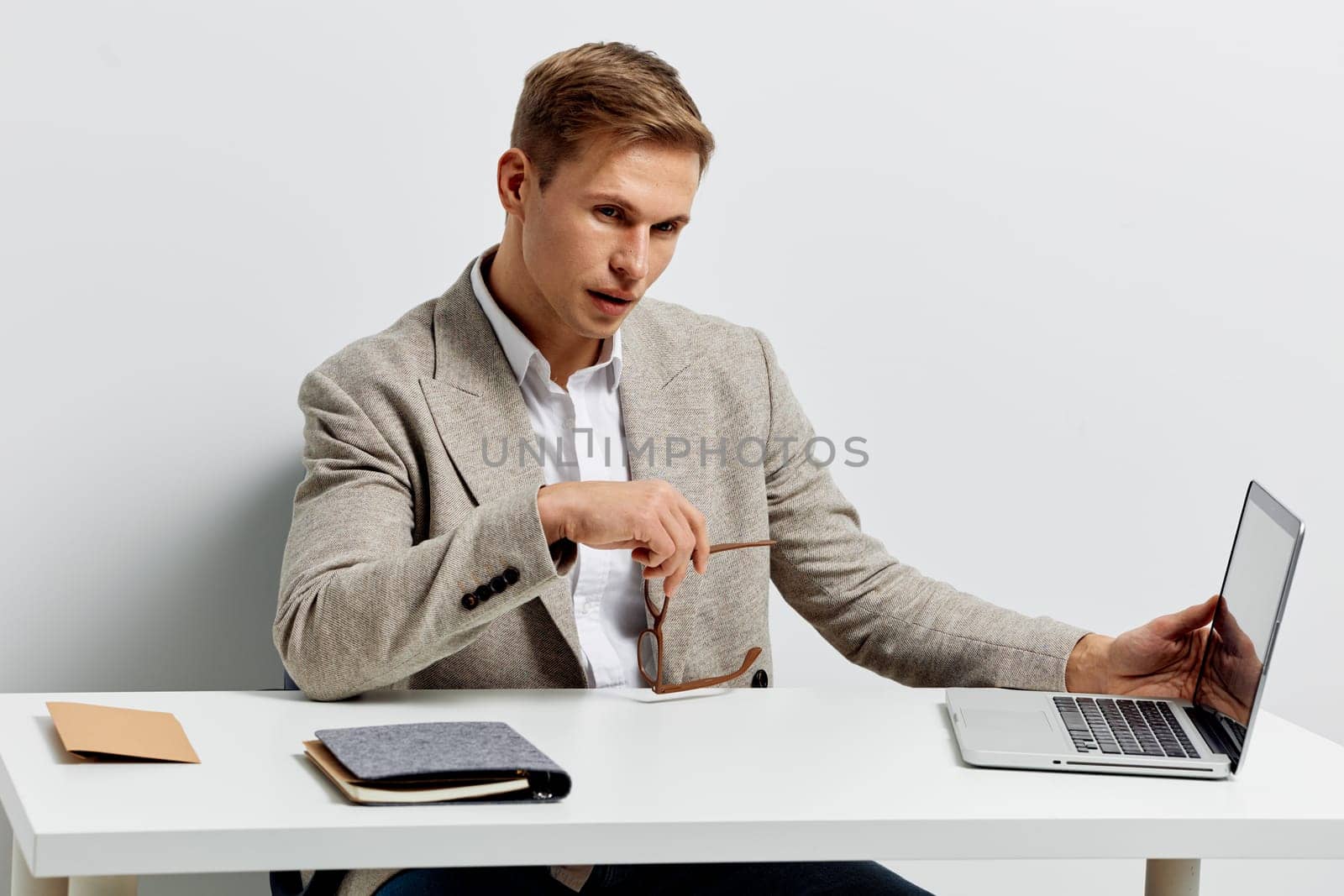 Focused young man with blonde hair dressed in a beige blazer working on a laptop and holding eyeglasses at a modern workspace, emphasizing productivity, technology, and business success. Concept: professional work environment