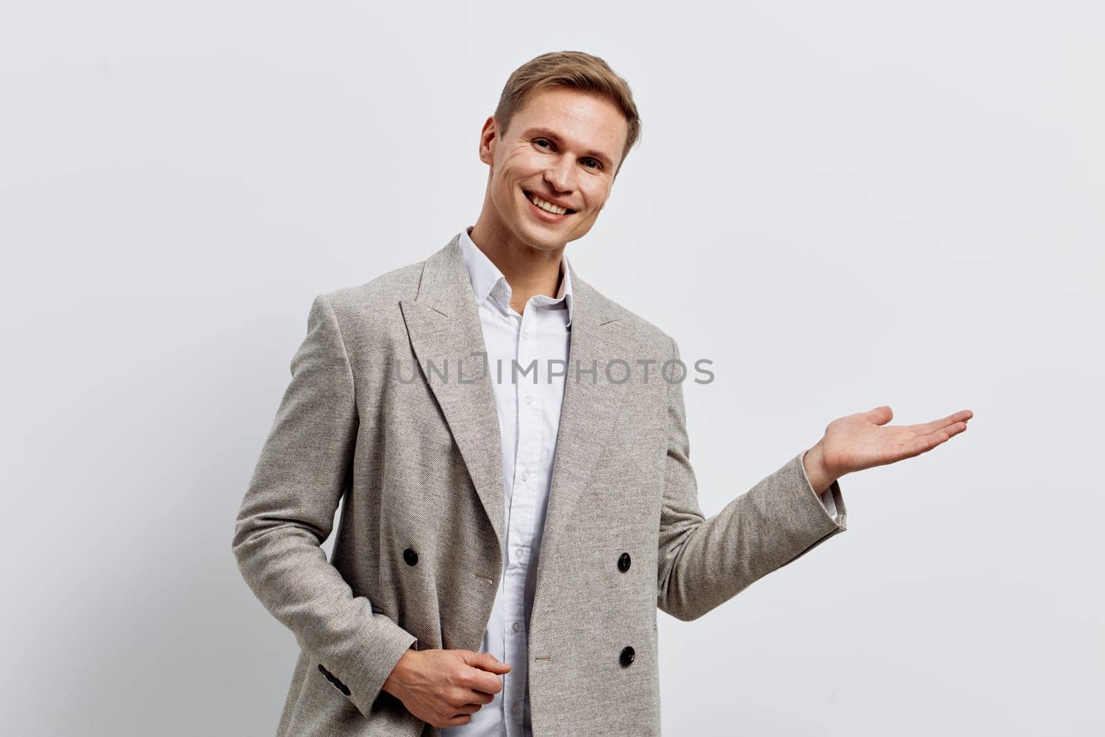 smiling man working digital nomad hold palm open wearing gray casual blazer and white shirt isolated on light background, confident cheerful young caucasian man representing remote work and modern technology lifestyle concept