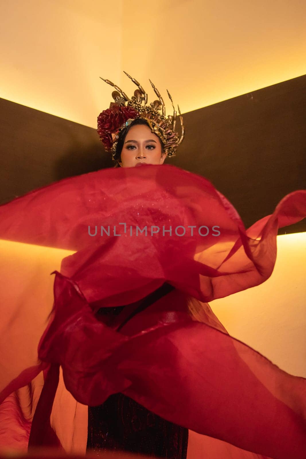 A glamor woman poses with a flying red dress on her body while wearing a gold crown inside the cream studio