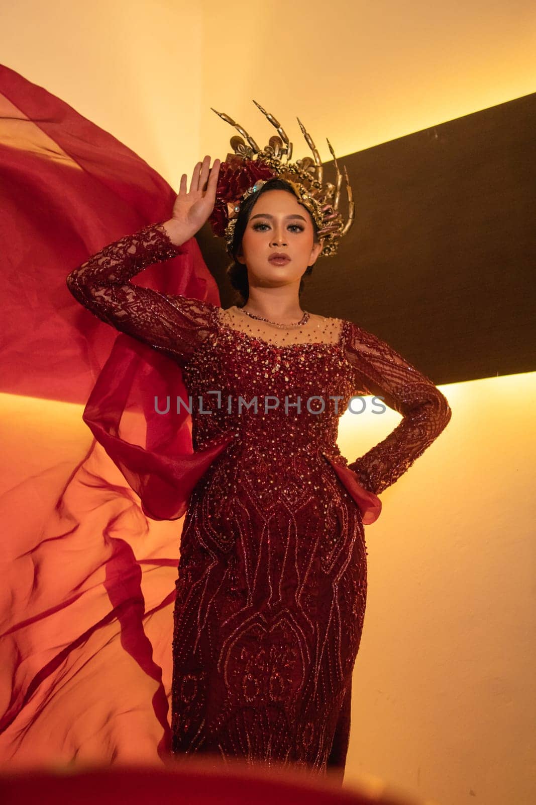 A glamor woman poses with a flying red dress on her body while wearing a gold crown inside the cream studio